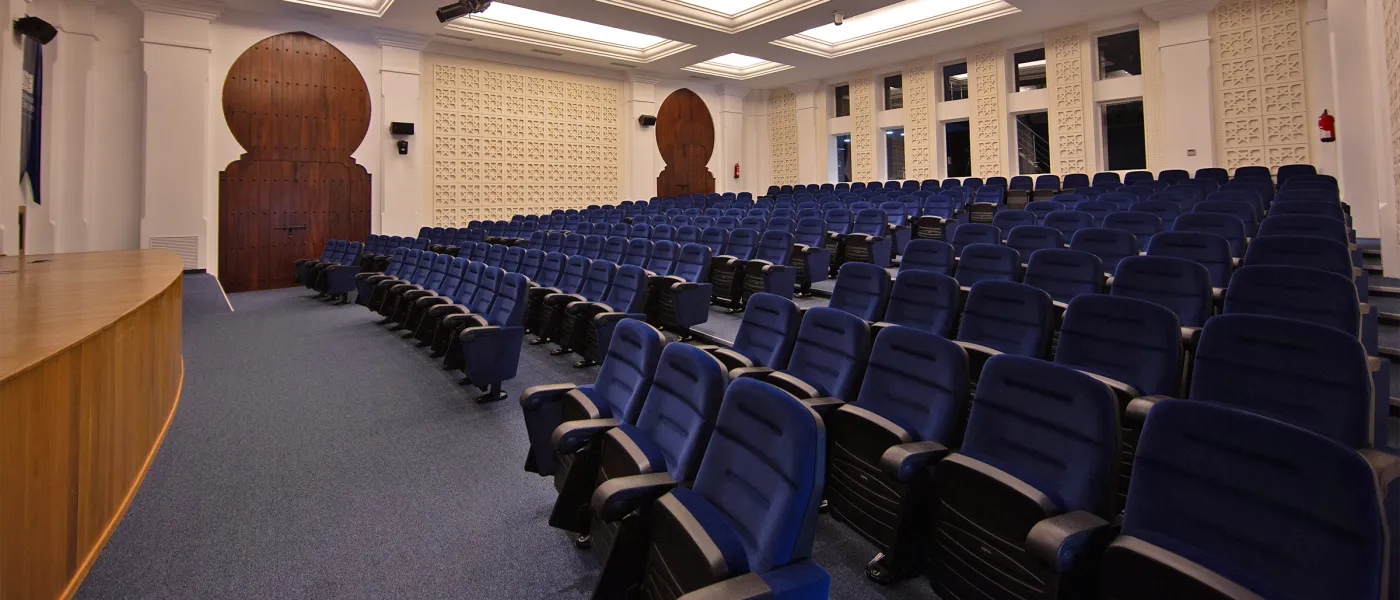 Auditorium with theater-style seating in navy blue, featuring white walls with decorative Moroccan carved panels and distinctive horseshoe-arched wooden doors.
