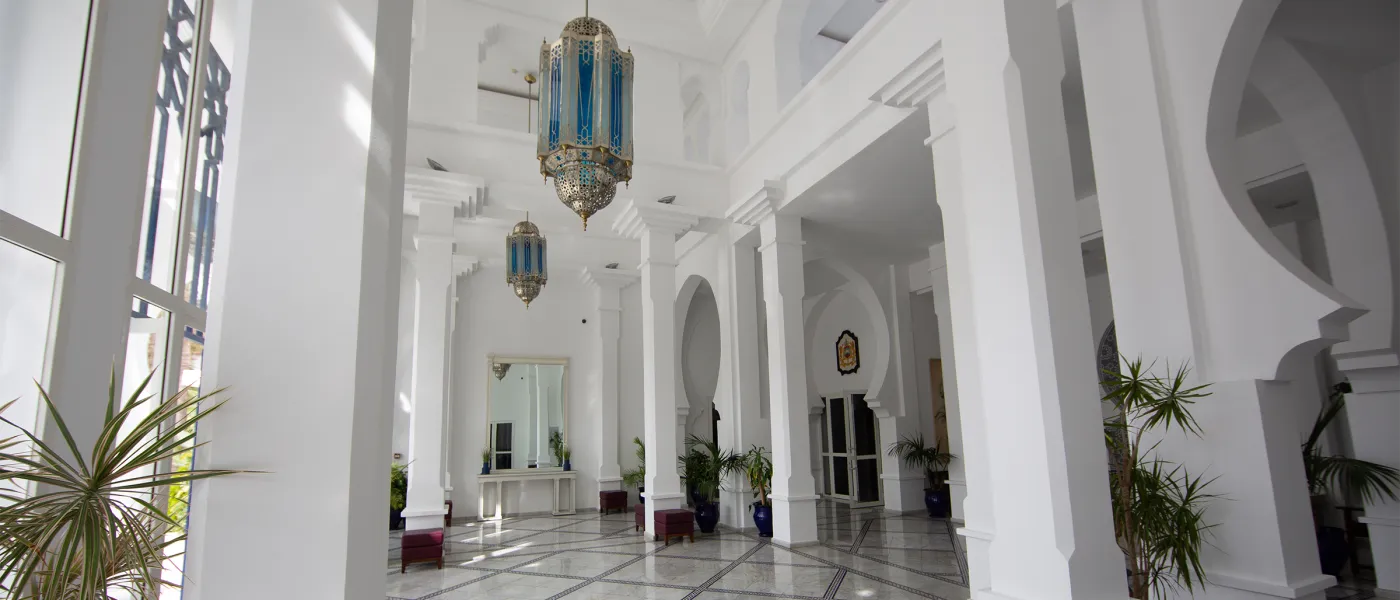 Bright white interior hallway with soaring ceilings, Moroccan arches, ornate blue and brass hanging lanterns, potted plants, and marble floors.