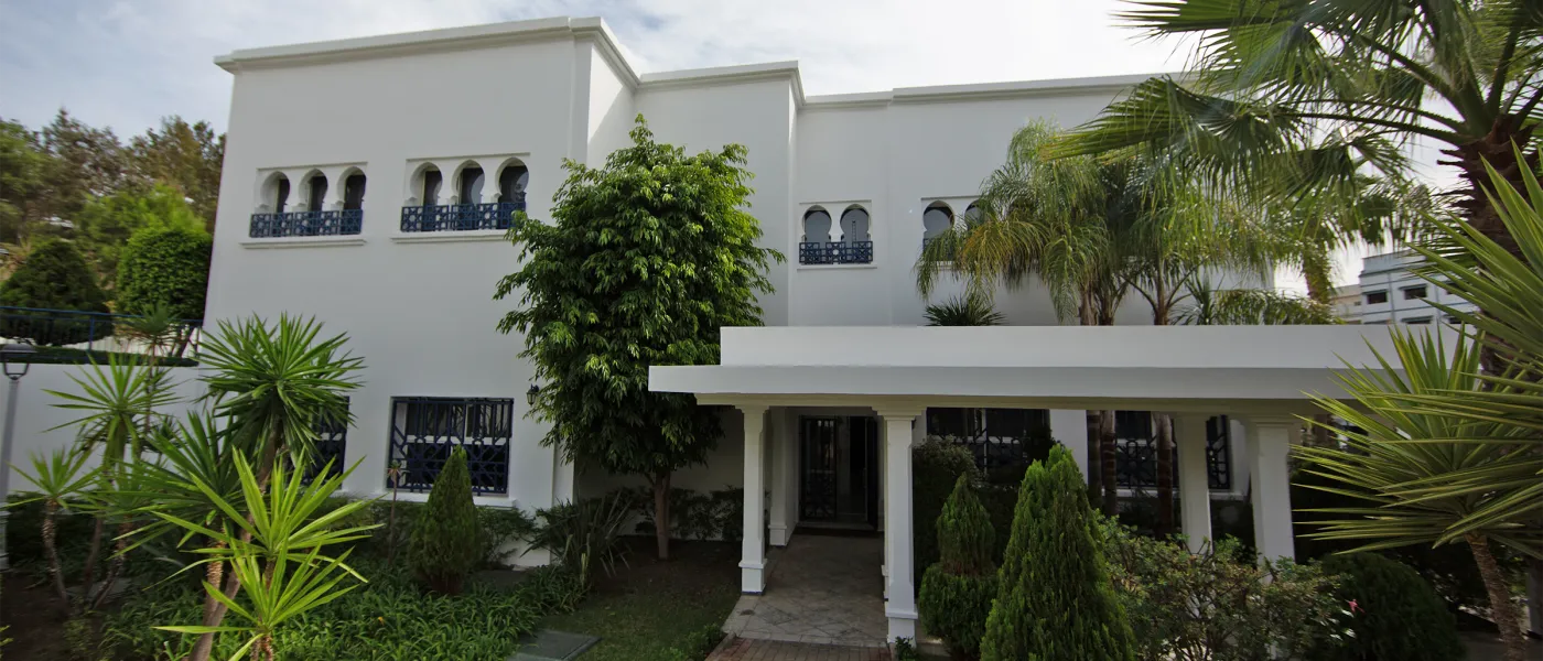 White Moroccan-style building with ornate balconies surrounded by lush tropical landscaping and palm trees at UNE Tangier Campus.