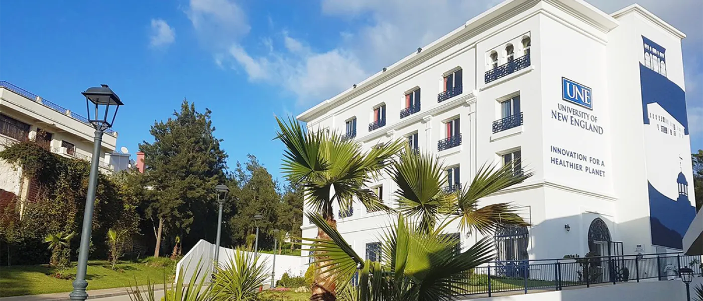 Campus building exterior with palm fronds in foreground, showing white facade with ornamental iron balconies and UNE branding reading "Innovation for a Healthier Planet."