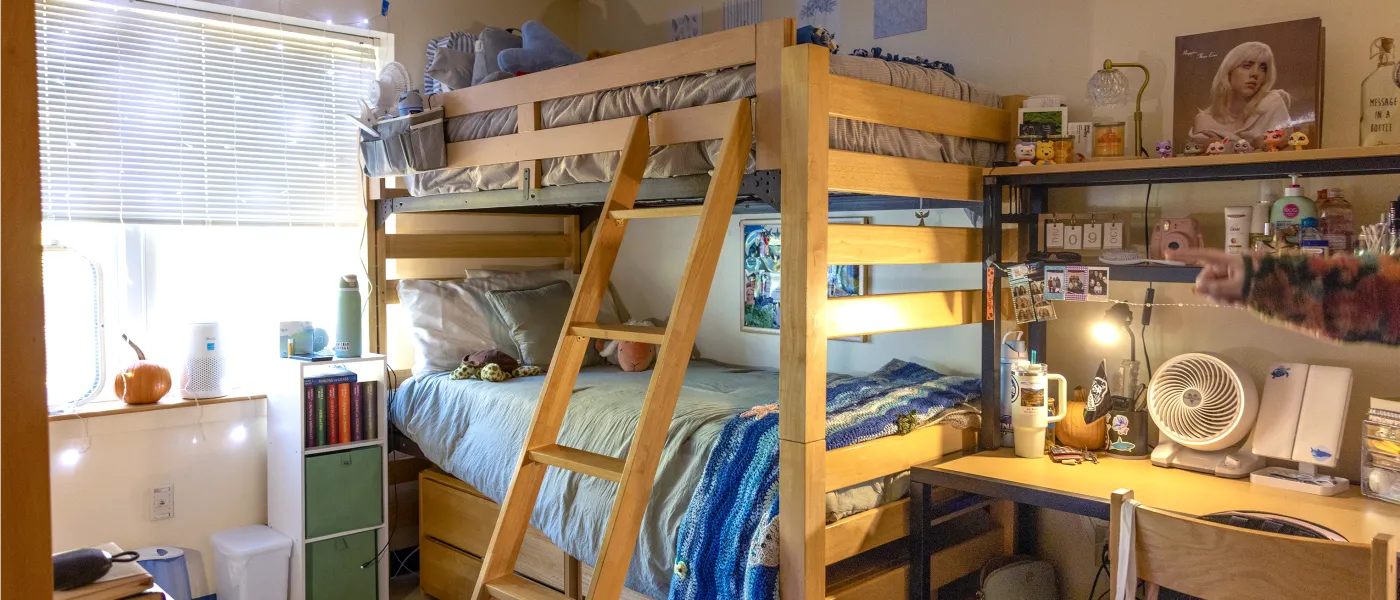 Dorm room with wooden lofted beds, built-in desk area with organized shelving, personal decorations including posters and photos, storage cubes, and a blue bedspread on the lower bunk.