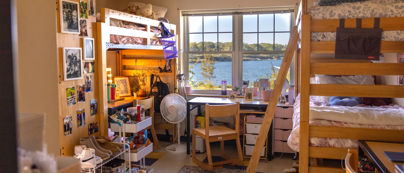 Dorm room with lofted beds on both sides, a desk and chair positioned between them facing a large window with a water view, personal items and photos displayed throughout.