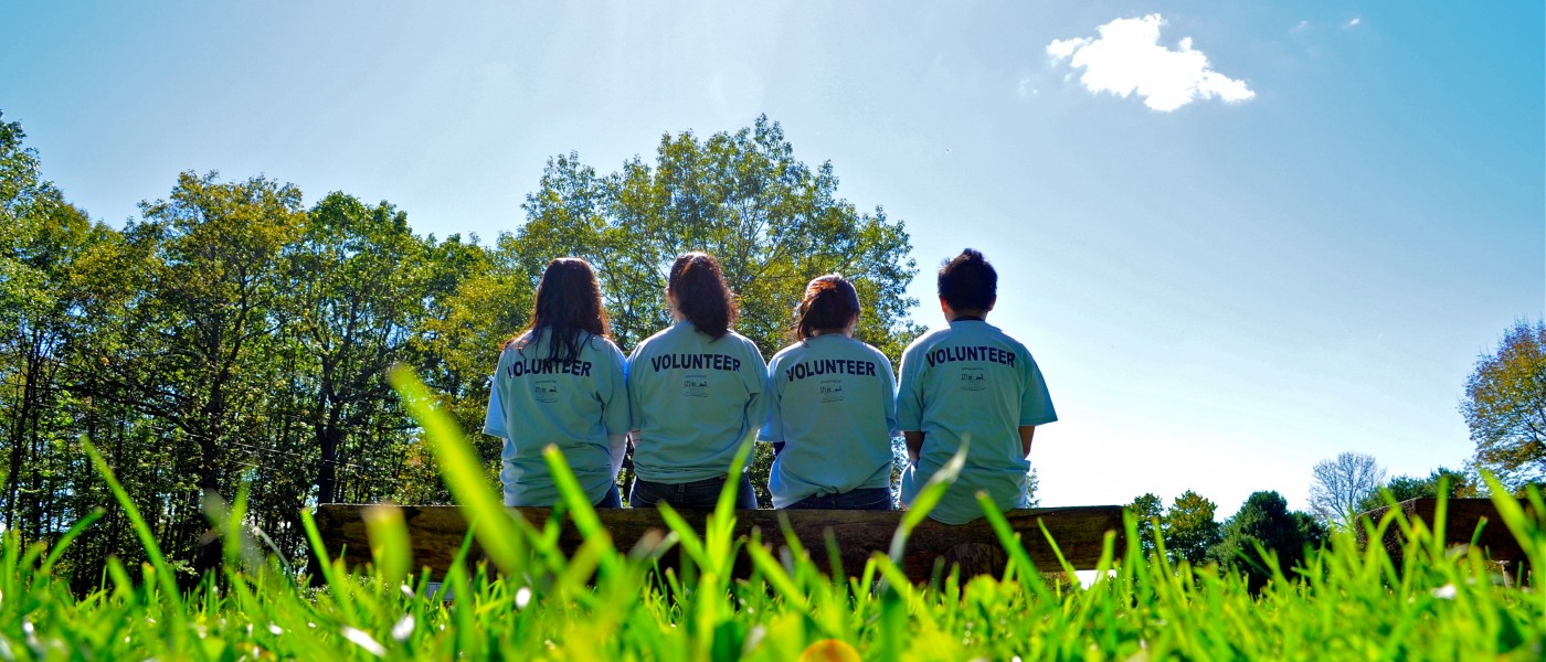 Four pharmacy students sit shoulder-to-shoulder on an outdoor bench wearing volunteer shirts