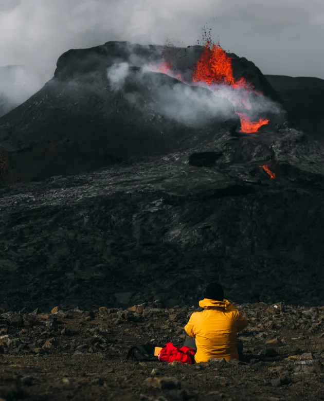 A person in a yellow winter jacket sits in front of an erupting volcano