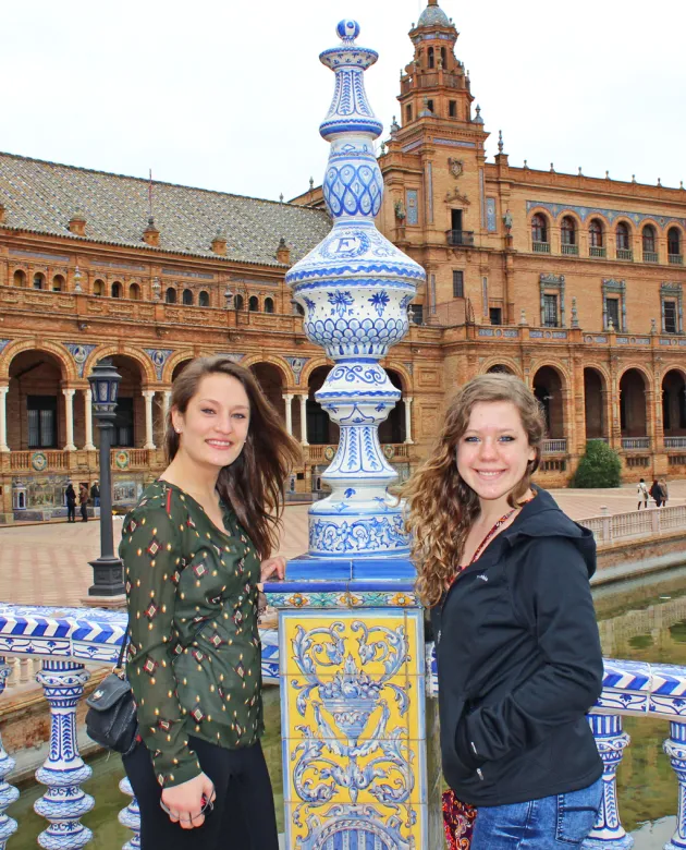 Two students stand with a decorative post on a bridge in front of a Spanish building