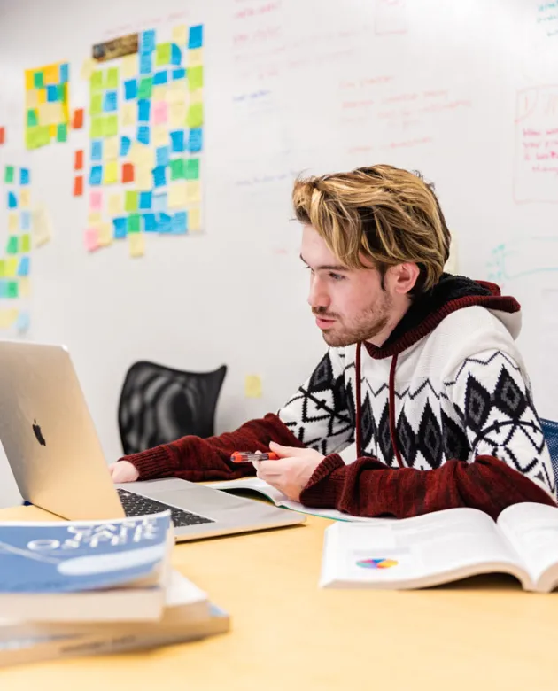 A U N E student studies on a laptop in front of a whiteboard covered in colorful post-it notes