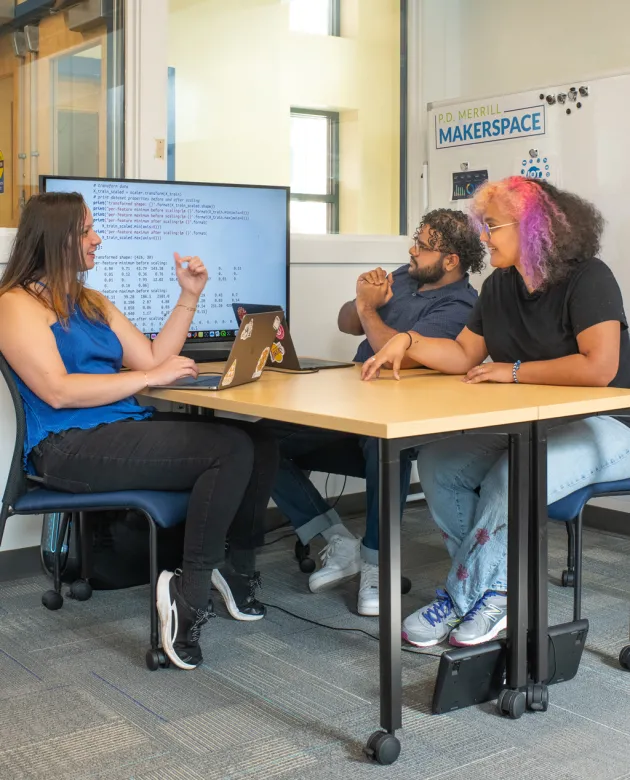 Three students sit in front of a large monitor that is displaying computer code