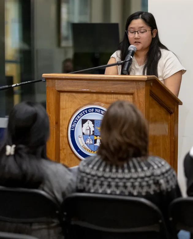 A student speaks from a podium to a crowd of other U N E students at a Creative Writing Club meeting
