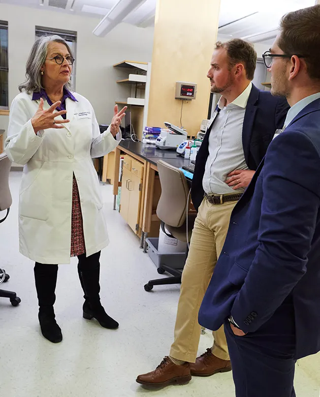 Karen Housenecht in a white coat in the lab speaks to two men in business attire. 