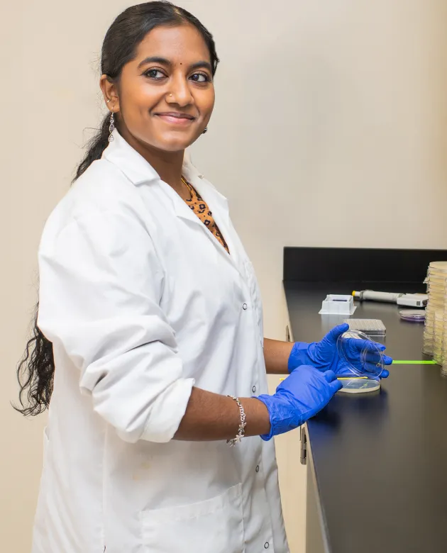 A UNE student in a white coat holds the lid to a petri dish at the counter of ther Burkholder lab while smiling toward the camera
