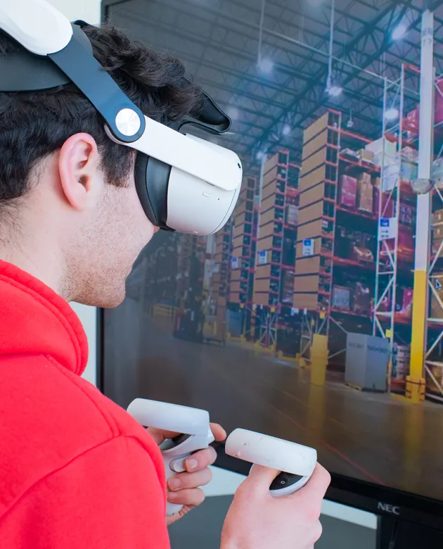 A student wears a V R headset and uses the controls to walk through a virtual warehouse