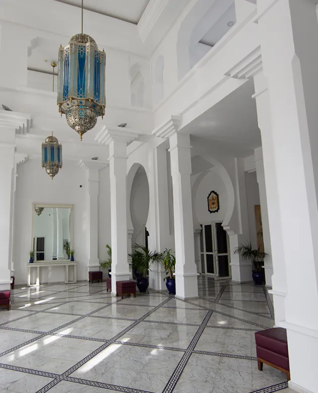 A large white hall featuring tall columns and an elegant chandelier hanging from the ceiling, with decorative molding and tiled flooring