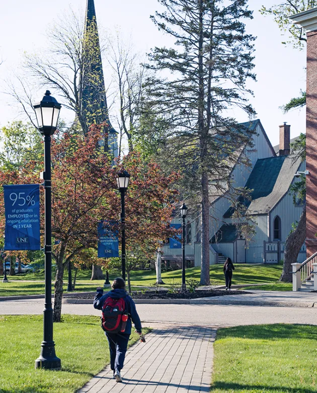 A student walks towards a building with a steeple from a paved path lined by green grass