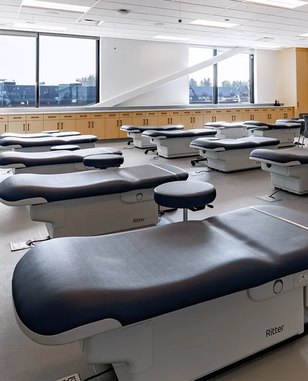A row of navy exam tables in front of a wall spanned by large windows and light-wood cabinets