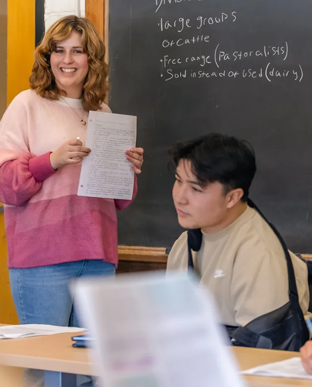 A student holding a piece of paper stands in front of a blackboard in a classroom with other students