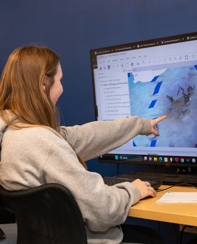 A student points at a large monitor displaying geographic mapping data, working at a desk with a blue wall behind them.