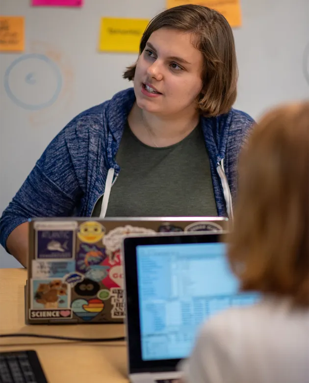 A student sits at a table during a collaborative session, with their sticker-covered laptop open and colorful sticky notes visible on the wall behind them. Another student's laptop is partially visible in the foreground.
