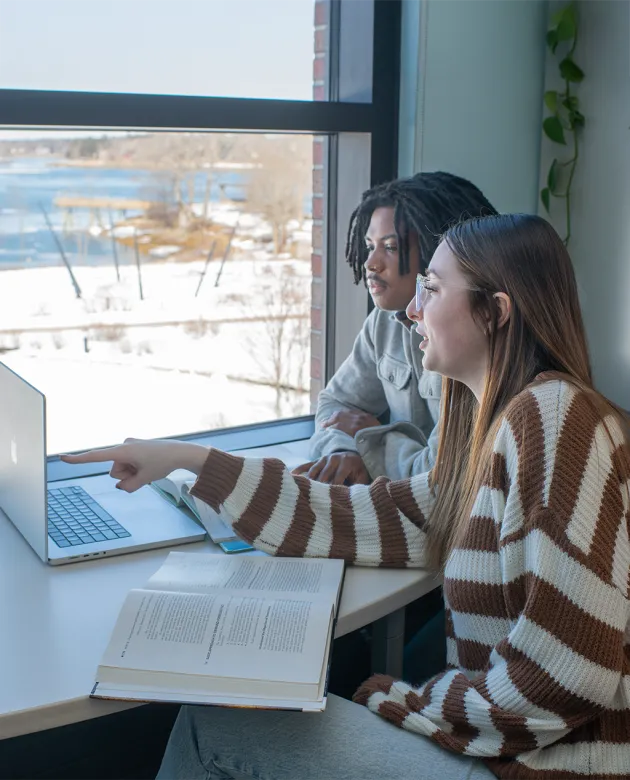 Two students collaborate at a desk by a window overlooking a snowy campus, working on a laptop with open textbooks beside them.