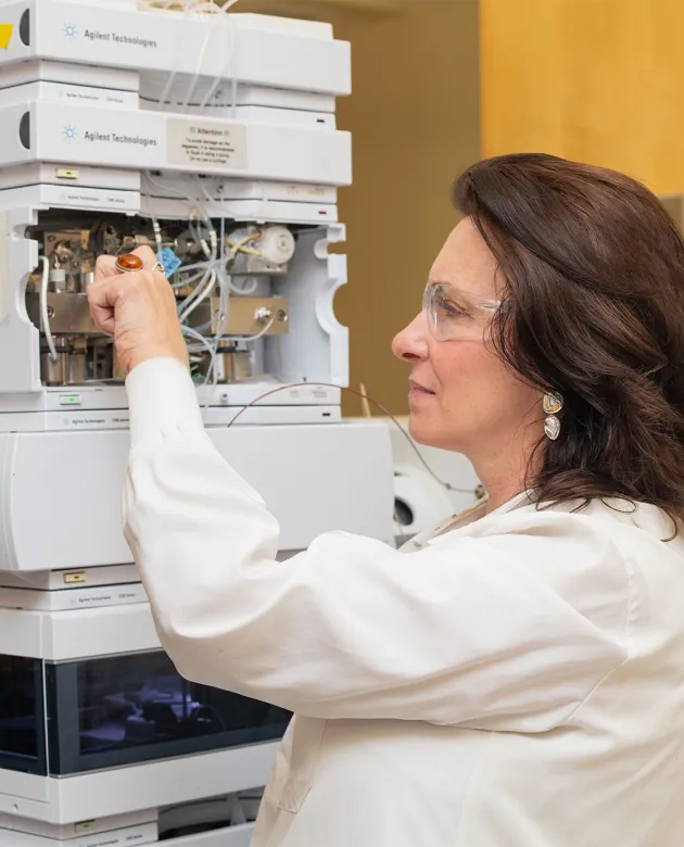 Deborah Barlow in a white lab coat and safety glasses examines a sample vial while working with Agilent Technologies laboratory equipment.