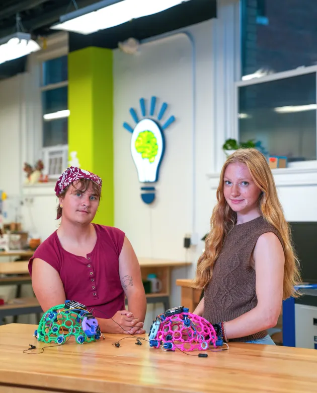 Milo Lypps and Jessica Howard pose with colorful skull caps in a makerspace with a lightbulb logo on the wall behind them.