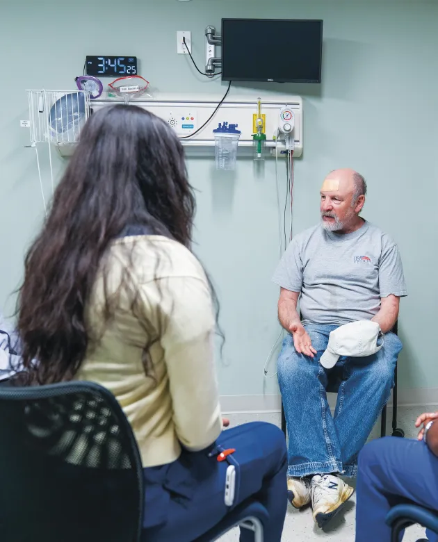 Michael, a patient actor, sits on a hospital chair addressing an interprofessional health care student team including nursing and dental medicine students during a clinical simulation.