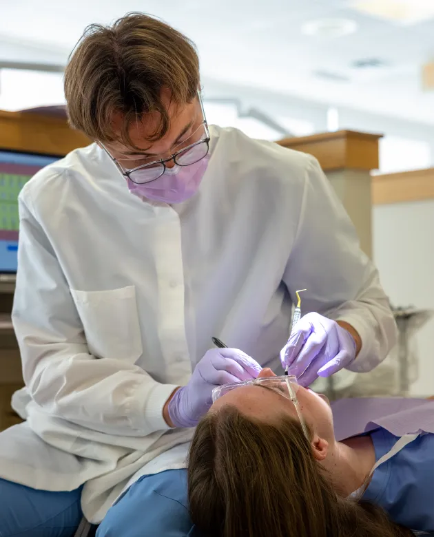 A dental hygiene student wearing purple gloves and a mask performs an oral examination on a patient reclined in a dental chair.