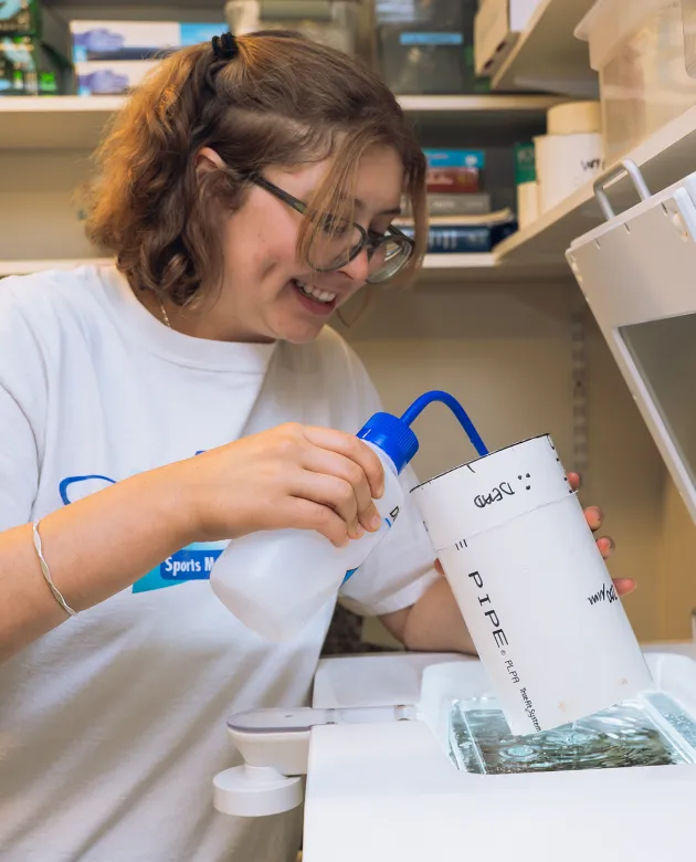 A graduate marine science student pours liquid from a bottle into a container while conducting laboratory research on plankton, with lab equipment visible in the background.