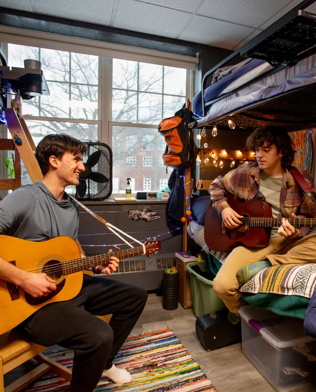 Two roommates sit across from each other playing acoustic guitars in their dorm room, decorated with string lights, a colorful rug, and personal belongings. A brick campus building is visible through the window.