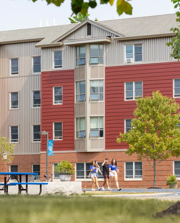 Three students walk past a modern four-story residence hall with red and tan horizontal siding over a brick base, framed by trees and outdoor seating areas.