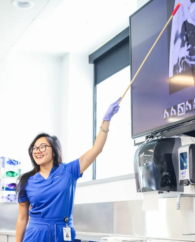 A UNE student in blue scrubs and a latex glove smiles while using a pointer to indicate a detail on a medical imaging scan displayed on a monitor in a clinical anatomy lab.