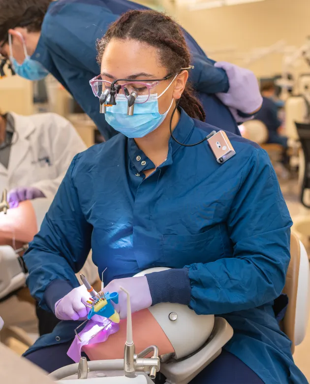 A UNE DMD student wearing loupes, a mask, and gloves practices a restorative dental procedure on a simulation model during a skills lab, with other students working in the background.