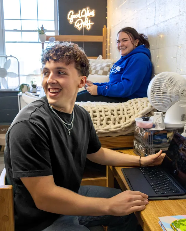 Two students laugh together in a decorated dorm room, one seated at a desk with a laptop and the other relaxing on a lofted bed.