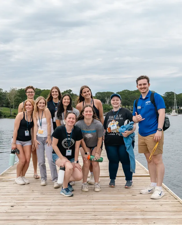 A group of new UNE students and an orientation leader pose together on a waterfront dock during a campus orientation event, with a sailboat and tree-lined cove visible behind them.