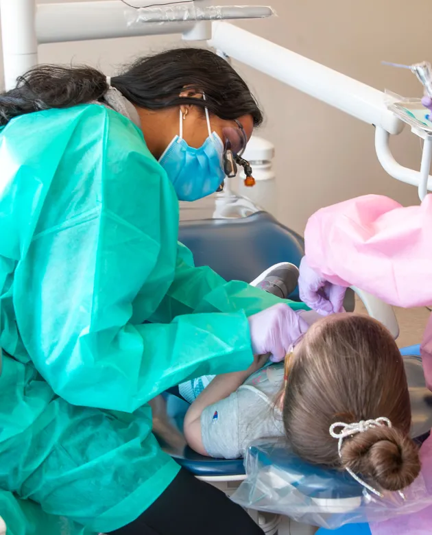 A U N E dental student in a mask and gown examines a young child's teeth in a dental chair at a community smile event.