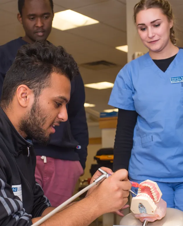 A high school student practices on a model of teeth using dental tools while a peer and dental hygiene student look on in a U N E Dental Sim Lab.