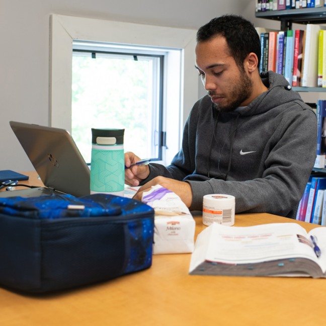 A male student studies in the library with textbooks and a laptop