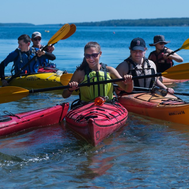 Five students and faculty members sit on the ocean in colorful kayaks