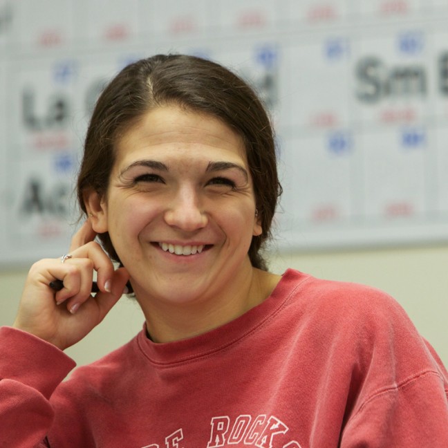 a student smiles in front of a poster of the periodic table