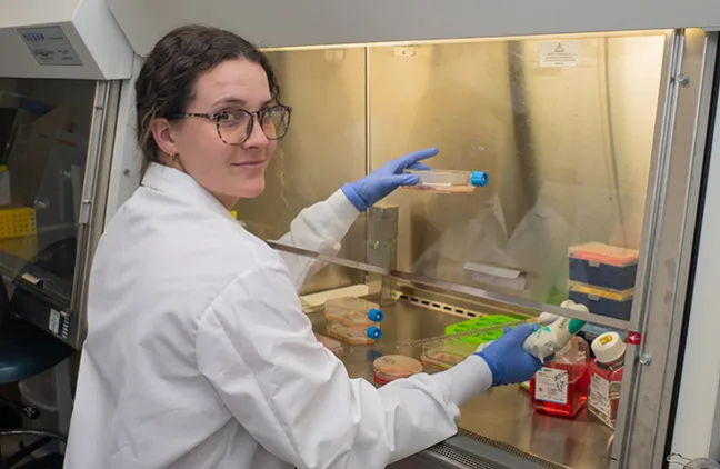 A student in a white lab coat smiles at the camera