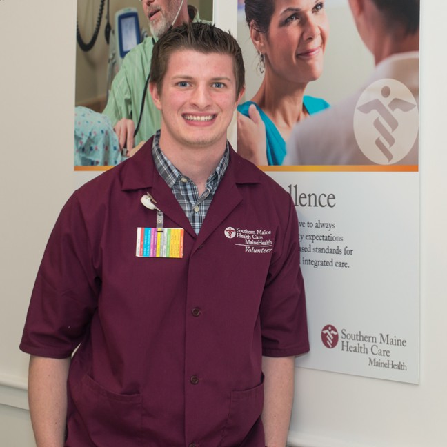 A U N E student wearing a colorful badge stands in front of a Southern Maine Health Care poster