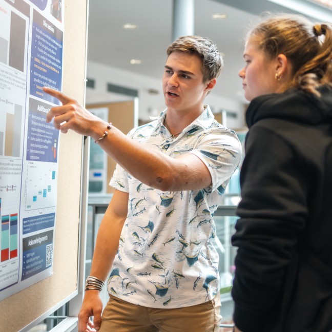 A student points to their presentation poster as they talk to a fellow student