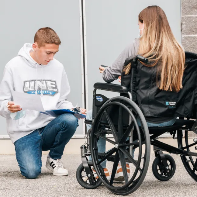 A student holding a clipboard kneels down next to another student in a wheelchair