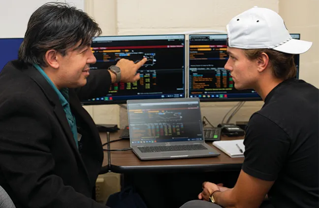 A business professor talks about finance to a student while pointing to dual monitors that display financial data