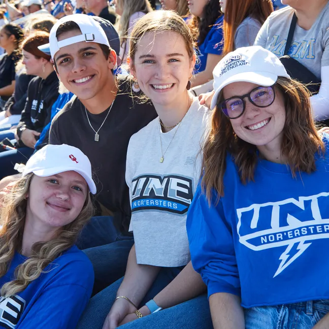 A group of four students in U N E t-shirts smile from a crowd in the stands of a football game