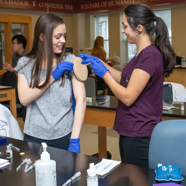 A pharmacy student practices placing a needle for administering a vaccine on another student during a lab