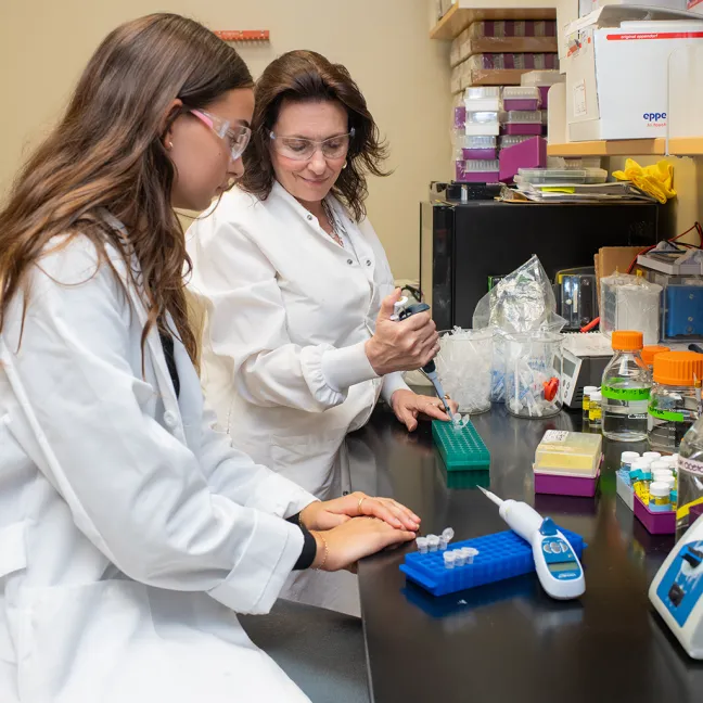 Two lab team members in safety glasses collaborating at a workbench, with one demonstrating multichannel pipette technique over sample plates while the other observes.