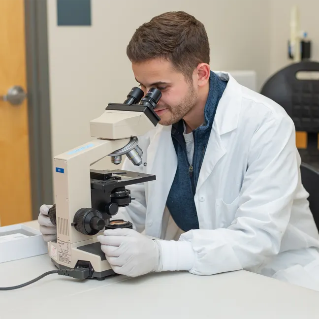 Lab technician in a white coat and gloves examining a slide specimen through a microscope at a laboratory workstation.