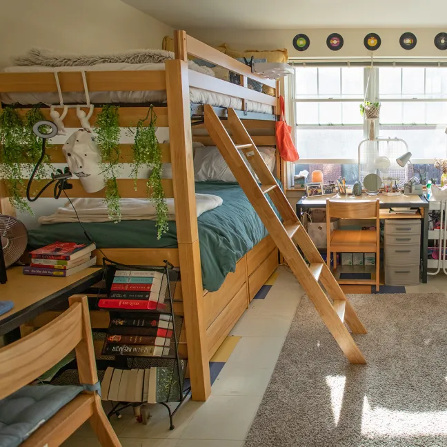 Bright dorm room with wooden lofted beds, hanging plants, a book cart, study desk near the window, and decorative vinyl records mounted on the wall.