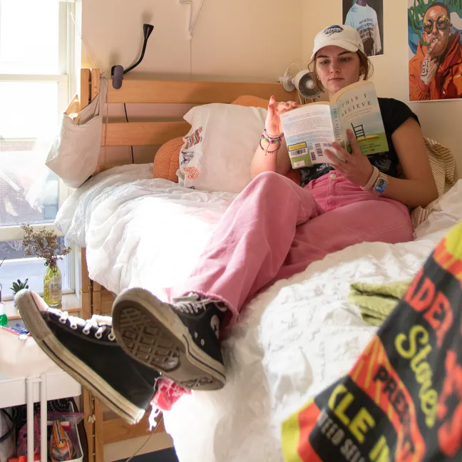 Bright dorm room with wooden lofted beds, hanging plants, a book cart, study desk near the window, and decorative vinyl records mounted on the wall.