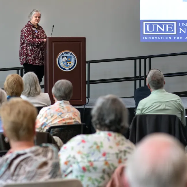 A speaker presents at a UNE podium before a seated audience of older adults during a Legacy Scholars event.